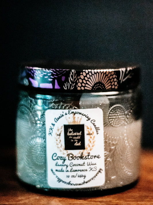 A close-up of the Cozy Bookstore candle in a glass jar in a refined ambiance with a label that says KK & Annie's Empowering Candles, She believed she could so she did, with a golden Queen's crown.