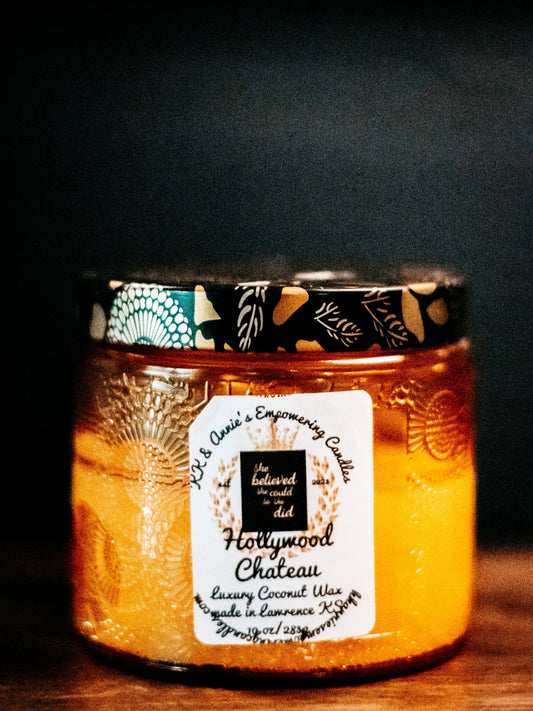 A close-up of the Hollywood Chateau candle in a glass jar in a refined ambiance with a label that says KK & Annie's Empowering Candles, She believed she could so she did, with a golden Queen's crown.