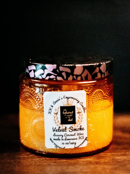 A close-up of the Velvet Smoke luxury candle in a glass jar in a refined ambiance with a label that says KK & Annie's Empowering Candles, She believed she could so she did, with a golden Queen's crown.