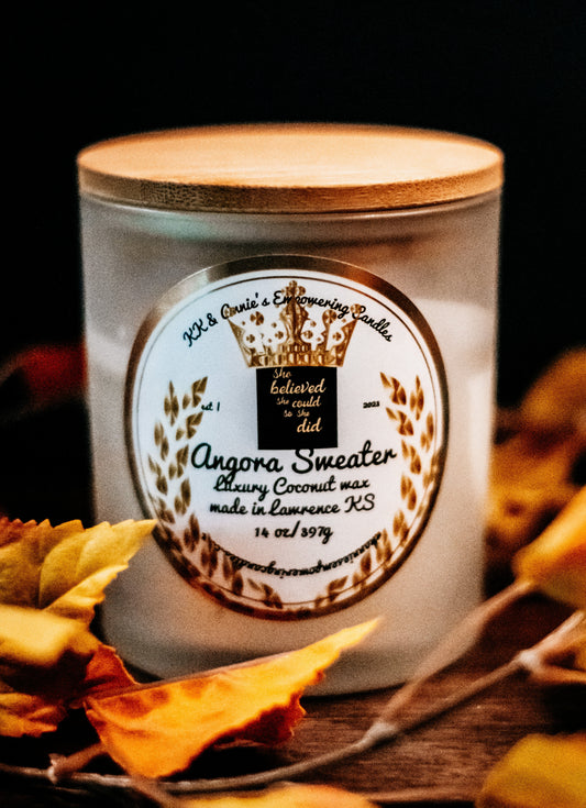 A close-up of the Angora Sweater candle in a glass jar in a refined ambiance with a label that says KK & Annie's Empowering Candles, She believed she could so she did, with a golden Queen's crown.