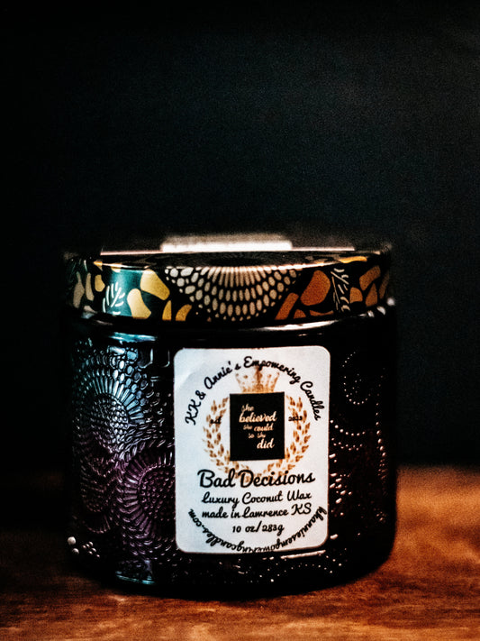A close-up of the Bad Decisions candle in a glass jar in a dimly lit bar setting with a label that says KK & Annie's Empowering Candles, She believed she could so she did, with a golden Queen's crown.