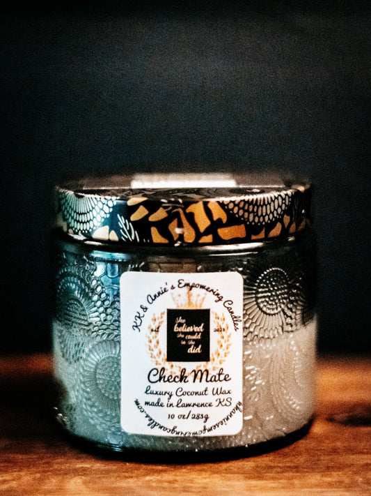A close-up of the Check Mate candle in a glass jar in a refined ambiance with a label that says KK & Annie's Empowering Candles, She believed she could so she did, with a golden Queen's crown.