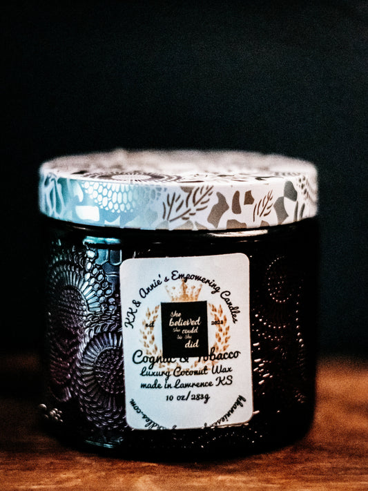 A close-up of the Cognac And Tobacco luxury candle in a glass jar in a refined ambiance with a label that says KK & Annie's Empowering Candles, She believed she could so she did, with a golden Queen's crown.