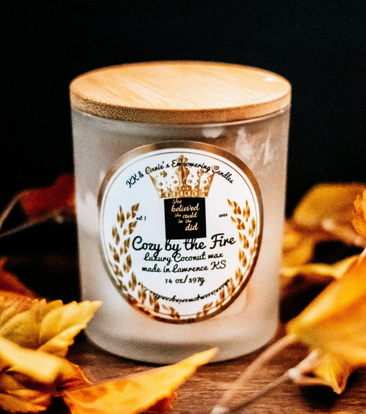 A close-up of the Cozy By The Fire candle in a glass jar in a refined ambiance with a label that says KK & Annie's Empowering Candles, She believed she could so she did, with a golden Queen's crown.