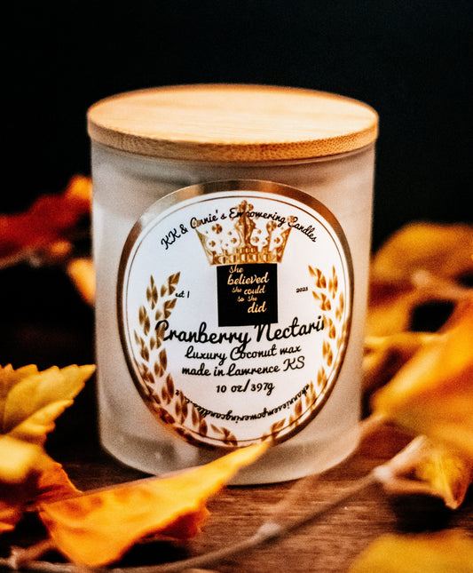 A close-up of the Cranberry Nectarine candle in a glass jar in a refined ambiance with a label that says KK & Annie's Empowering Candles, She believed she could so she did, with a golden Queen's crown.