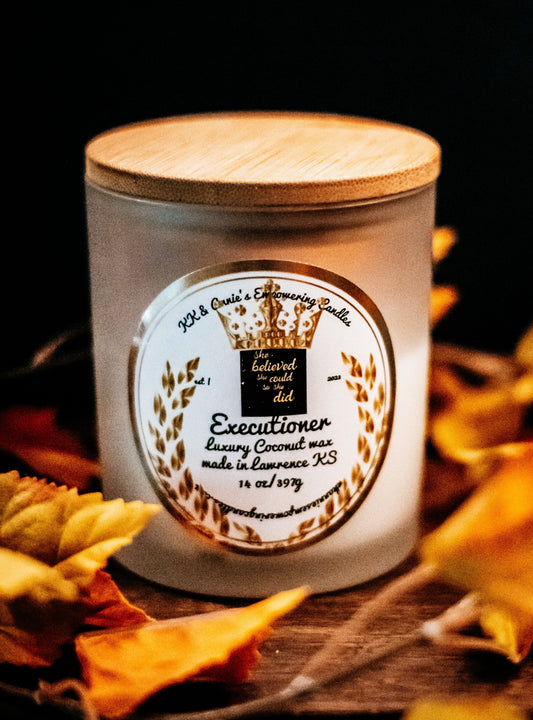A close-up of the Executioner candle in a glass jar in a refined ambiance with a label that says KK & Annie's Empowering Candles, She believed she could so she did, with a golden Queen's crown.