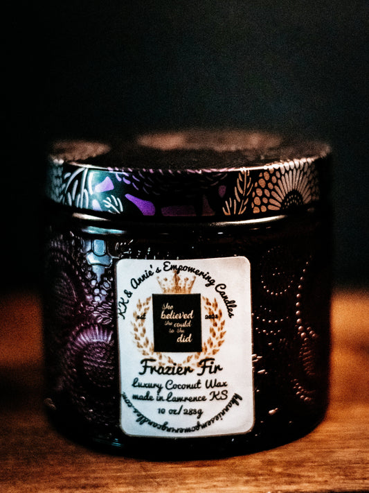 A close-up of the Frazier Fir luxury candle in a glass jar in a refined ambiance with a label that says KK & Annie's Empowering Candles, She believed she could so she did, with a golden Queen's crown.