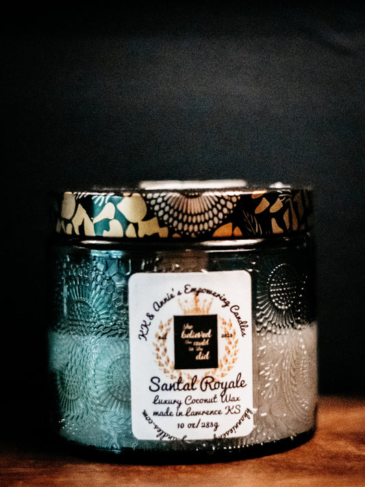 A close-up of the Santal Royale candle in a glass jar in a refined ambiance with a label that says KK & Annie's Empowering Candles, She believed she could so she did, with a golden Queen's crown.