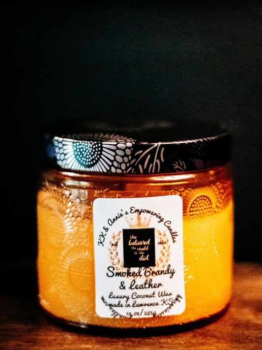 A close-up of the Smoked Brandy & Leather candle in a glass jar in a refined ambiance with a label that says KK & Annie's Empowering Candles, She believed she could so she did, with a golden Queen's crown.