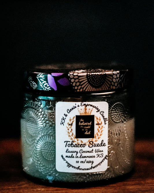 A close-up of the Tobacco Suede candle in a glass jar in a refined ambiance with a label that says KK & Annie's Empowering Candles, She believed she could so she did, with a golden Queen's crown.
