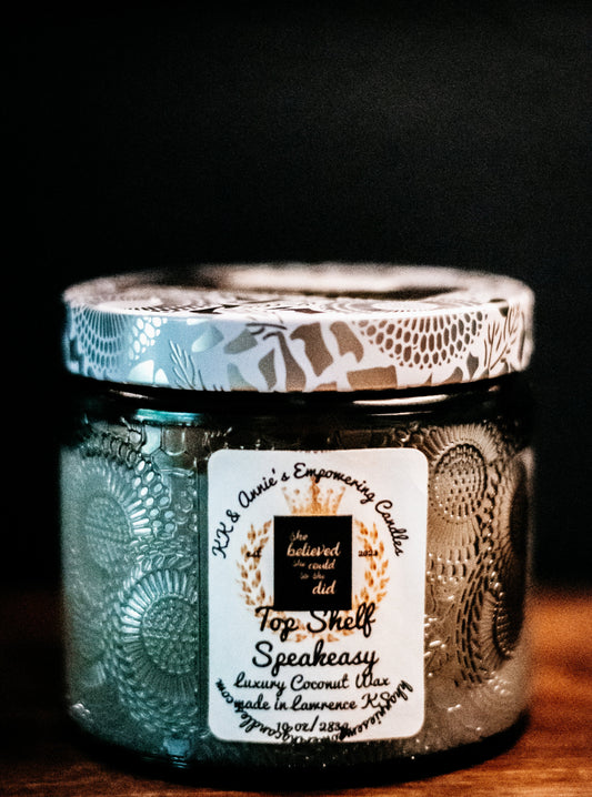 A close-up of the Top Shelf Speakeasy candle in a glass jar in a refined ambiance with a label that says KK & Annie's Empowering Candles, She believed she could so she did, with a golden Queen's crown.