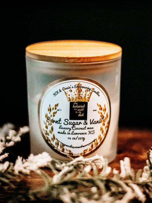 A close-up of the Burnt Sugar & Vanilla Bean candle in a glass jar in a refined ambiance with a label that says KK & Annie's Empowering Candles, She believed she could so she did, with a golden Queen's crown.