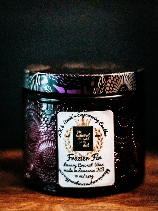 A close-up of the Deck the Halls festive candle in a glass jar in a refined ambiance with a label that says KK & Annie's Empowering Candles, She believed she could so she did, with a golden Queen's crown.